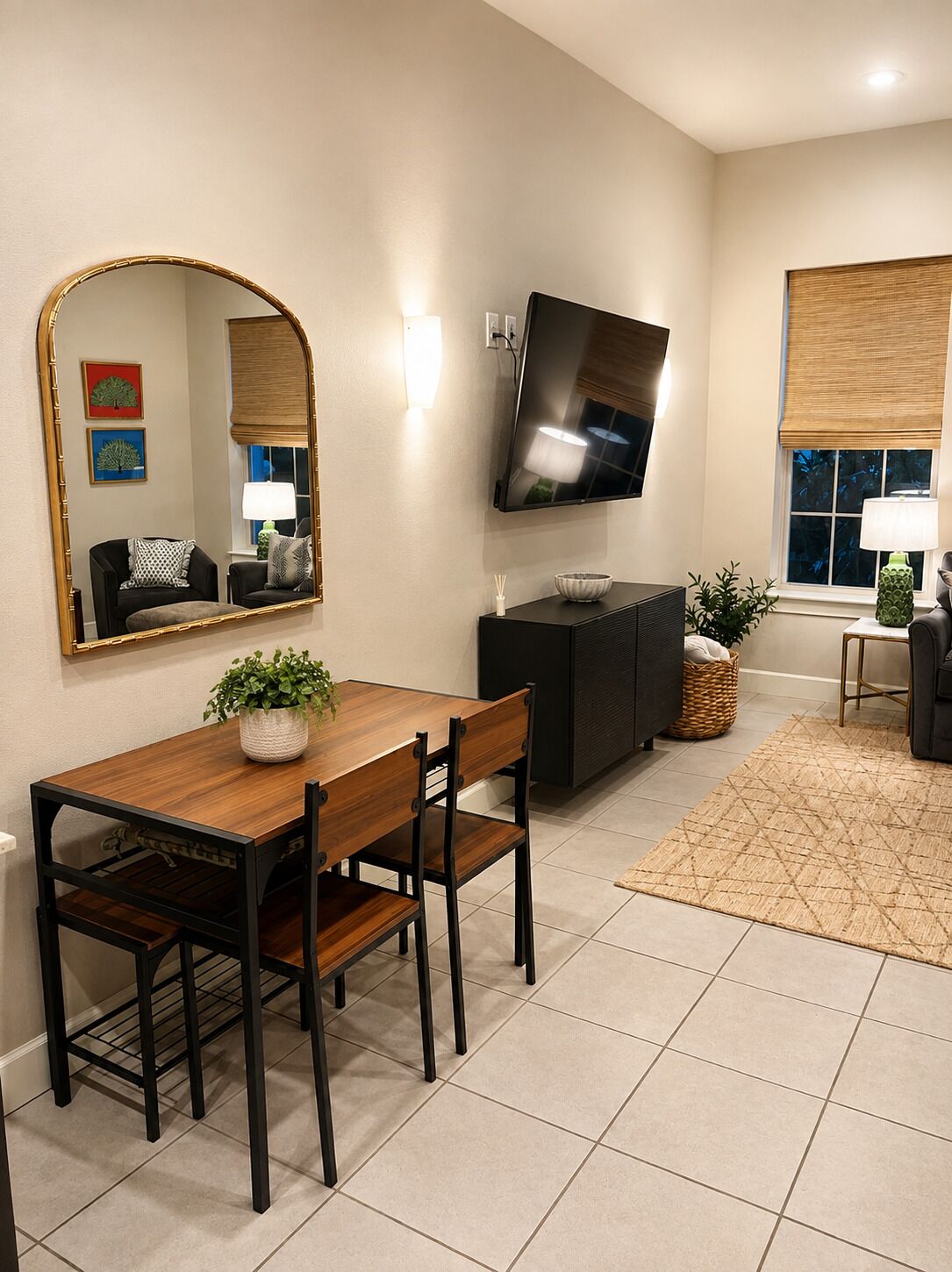 A modern living room with a dining table, chairs, a flat-screen TV mounted on the wall, and a large mirror.