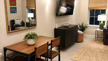 A modern living room with a dining table, chairs, a flat-screen TV mounted on the wall, and a large mirror.
