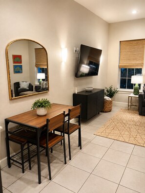 A modern living room with a dining table, chairs, a flat-screen TV mounted on the wall, and a large mirror. - Modern Downtown Bentonville Condo - Walk to It All! (Bentonville)