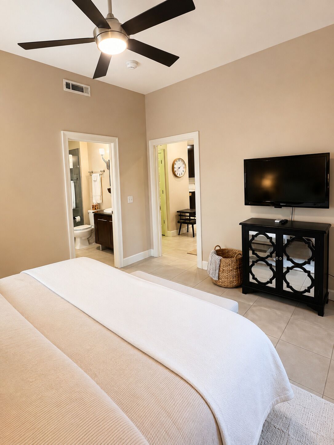 Ein Schlafzimmer mit Bett, Deckenventilator, Fernseher und einem durch eine offene Tür einsehbaren Badezimmer.