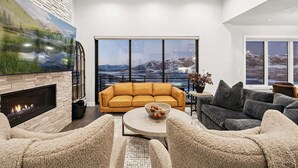 Ett modernt vardagsrum med en öppen spis, ett stort fönster med utsikt över bergen, en gul soffa och en grå soffa.