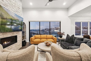 A modern living room with a fireplace, a large window with a mountain view, a yellow sofa, and a gray sofa. - MOUNTAIN LUXE | Panoramic Views + Private Hot Tub (Hideout)
