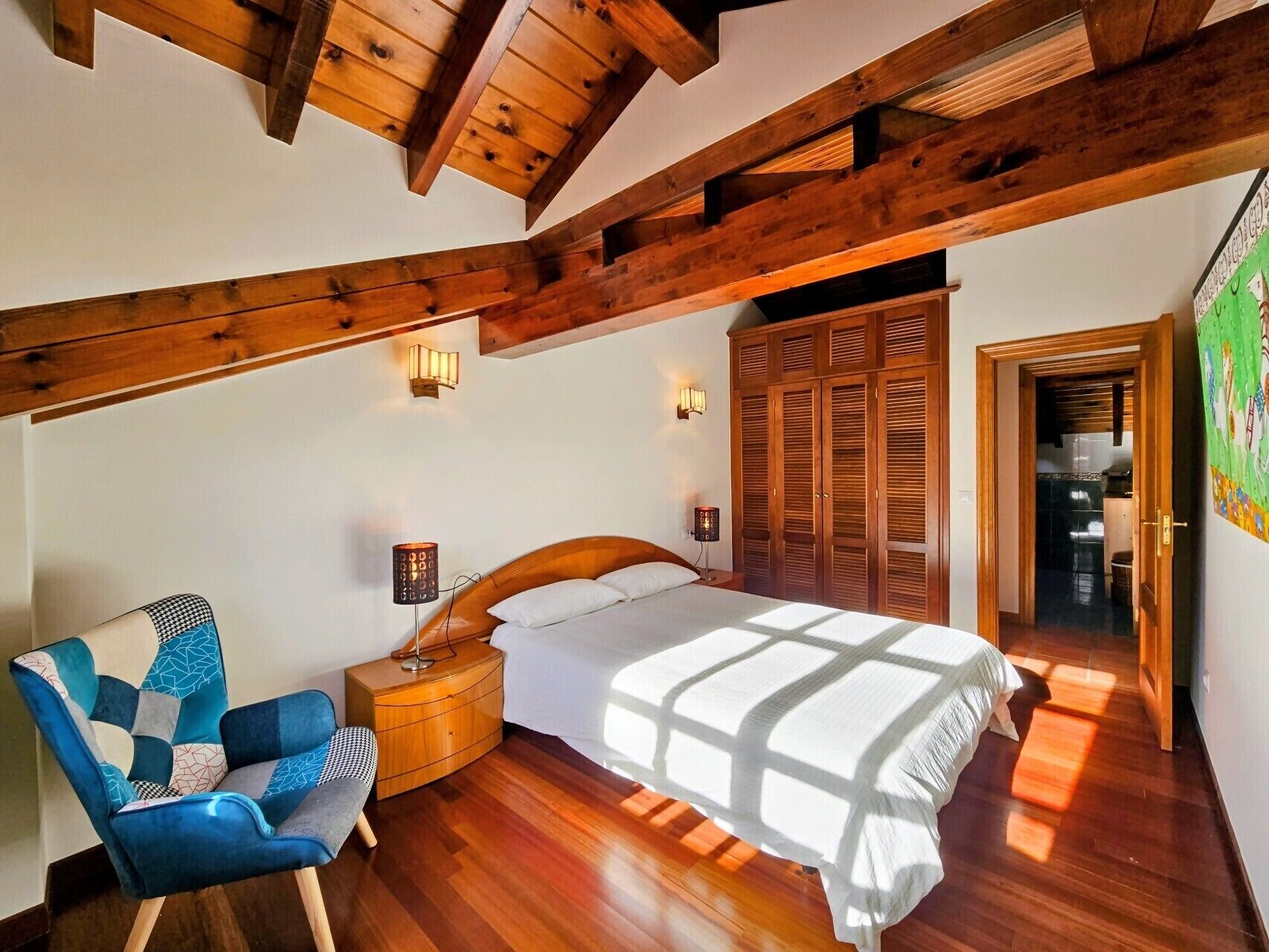 A bedroom with a wooden ceiling, a bed with a white bedspread, a blue armchair, and a wardrobe.