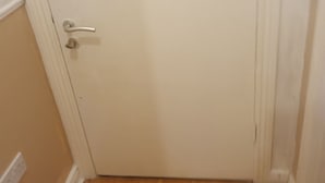 A closed white door with a silver handle and a keyhole, wooden flooring, and a white wall with a baseboard. - Cheap modern room (Dublin)