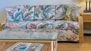 A sofa with floral upholstery, a glass coffee table with magazines, and a framed painting on the wall. - Bright condo in beautiful Honolulu with cool AC breeze (Honolulu)