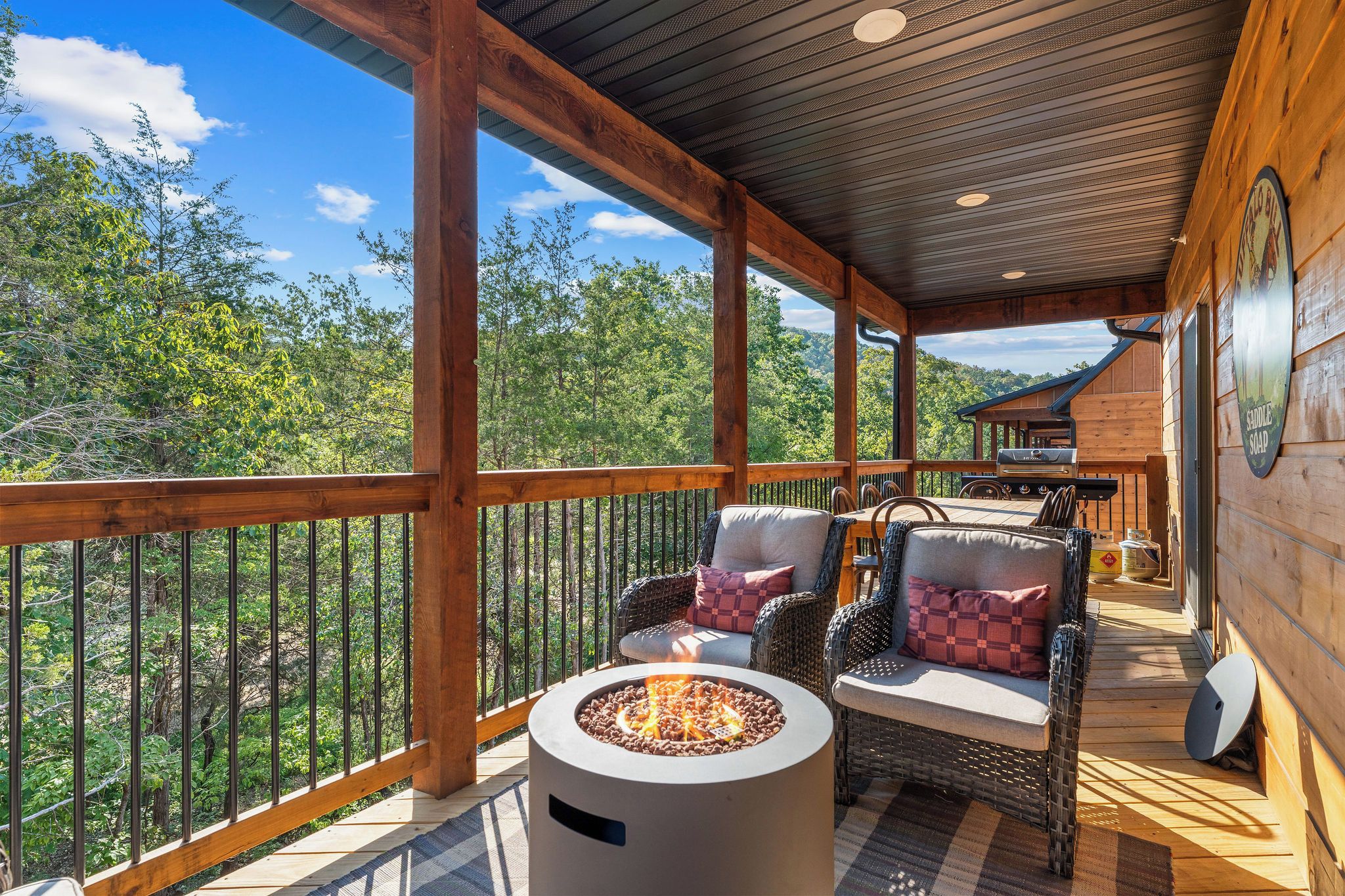 A covered outdoor deck with a fire pit, wicker furniture, and a view of the surrounding forest.