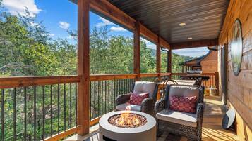 A covered outdoor deck with a fire pit, wicker furniture, and a view of the surrounding forest.