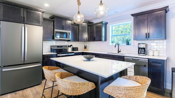 Una cocina moderna con armarios oscuros, electrodomésticos de acero inoxidable, una isla central con taburetes altos e iluminación colgante.