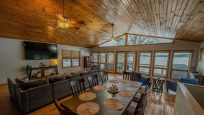 Amplia sala de estar con techo de madera, comedor con mesa larga y sillas, y ventanas grandes que ofrecen vistas a un lago.