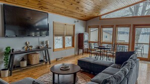 Una sala de estar con un televisor grande, techo de madera y ventanas amplias.