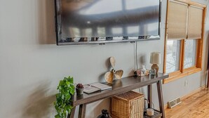 Una sala moderna con un televisor de pantalla plana grande montado en la pared, suelo y techo de madera, una ventana con persianas y una pequeña planta sobre una mesa consola.