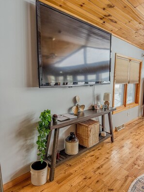 A modern living room with a large flat-screen TV mounted on the wall, wooden flooring, a wooden ceiling, a window with blinds, and a small plant on a console table.