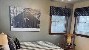 Een slaapkamer met een bed, een stoel, een lamp, een raam met jaloezieën, een plafondventilator en een aan de muur hangend schilderij.