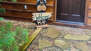 Une cabane en bois avec une porte brun foncé, une pancarte d’accueil ornée d’une découpe d’ours, et un chemin de pierres menant à l’entrée.