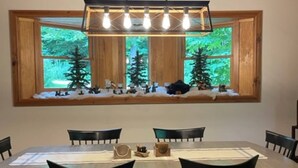 Un espace repas avec une grande table en bois, des chaises noires et une vue sur les arbres, visible à travers une fenêtre située au-dessus de la table.