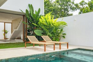 A modern outdoor pool area with two lounge chairs, surrounded by lush greenery and a white wall. - Tropical Modern Villa Amaya by OriVista (Pecatu)
