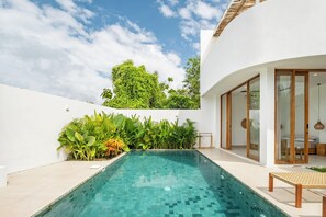 A modern outdoor pool area with a clear blue pool, surrounded by lush greenery and a white building with large glass doors. - Tropical Modern Villa Amaya by OriVista (Pecatu)
