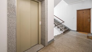 Un couloir avec un ascenseur de couleur beige, une porte en bois et un escalier doté d’une rampe en métal.
