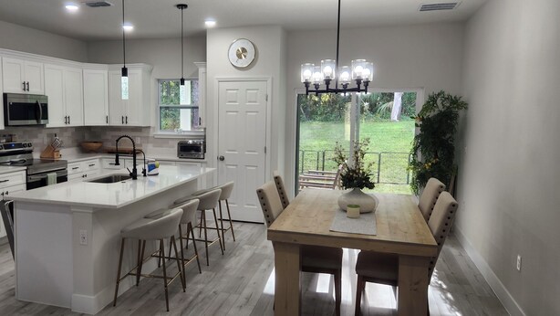 A modern kitchen with white cabinetry, stainless steel appliances, a central island with bar stools, and a dining area with a wooden table and chairs. - Beautiful New Home in Citrus Springs, sleep 6 (Citrus Springs)