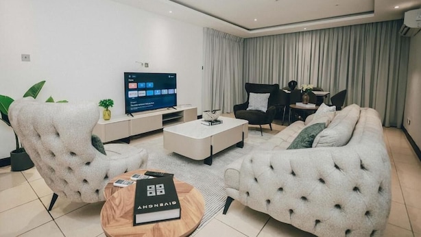 A modern living room with a flat-screen TV, two sofas, a coffee table, and a dining area with chairs. - Luxury 3BR Alema Court-Airport Close (Accra)