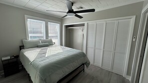 Une chambre à coucher comprenant un lit, un ventilateur de plafond, un dressing et une fenêtre avec des stores.