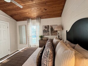 A bedroom with a wooden ceiling, a bed with patterned bedding, a nightstand with a lamp, and a window with curtains. - Charming 2-bedroom house in welcoming Waco TX (Waco)