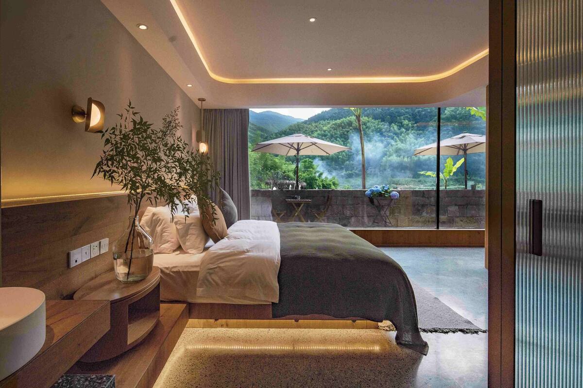 Un dormitorio moderno con una cama grande, una mesita de noche con un florero, vistas a un jardín exuberante a través de amplios ventanales, y un balcón con sombrillas.