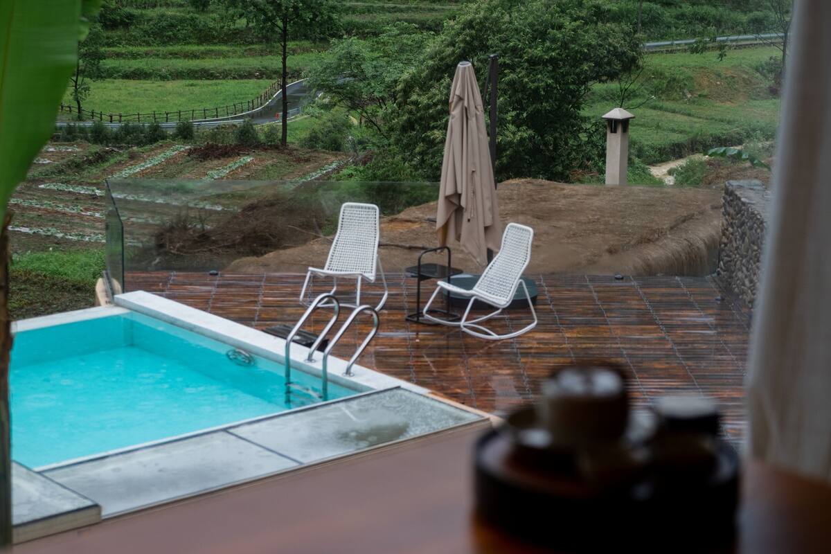 Una piscina con dos sillas reclinables blancas y una sombrilla, rodeada de una terraza de madera y vistas a campos escalonados.