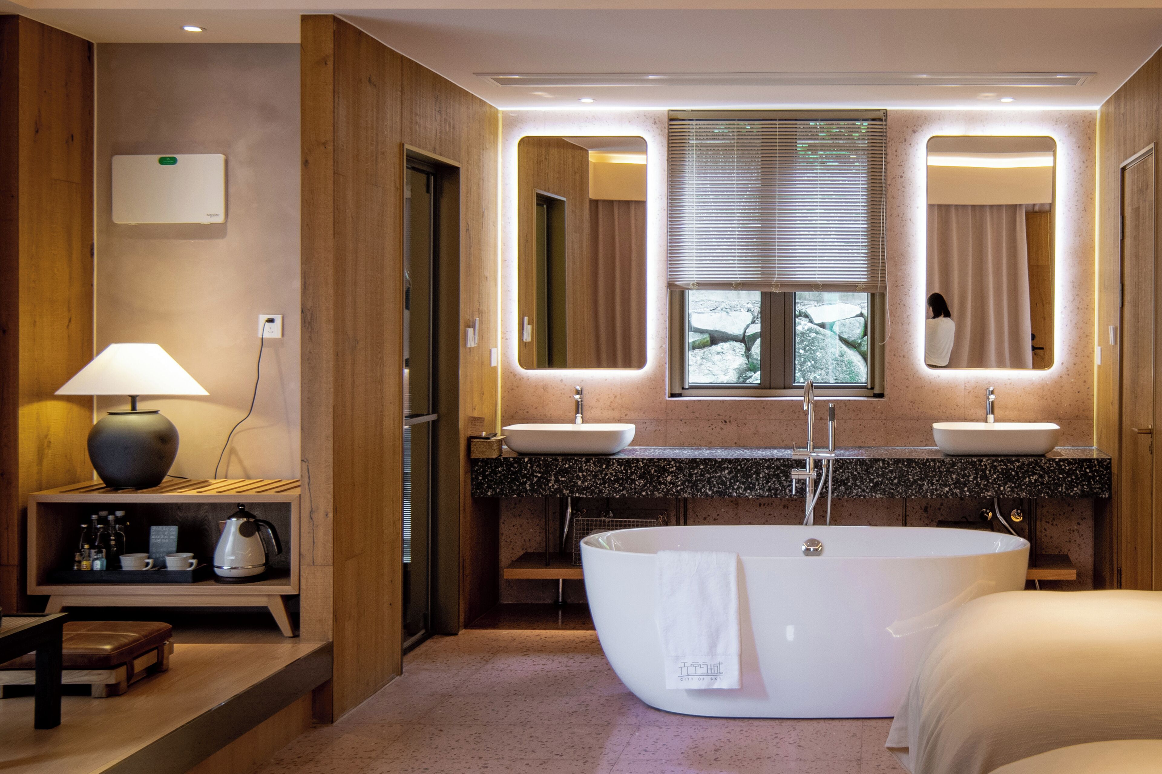 Ein modernes Badezimmer mit freistehender Badewanne, zwei Waschbecken, einem Waschtisch mit Spiegel und einem Fenster mit Jalousien.