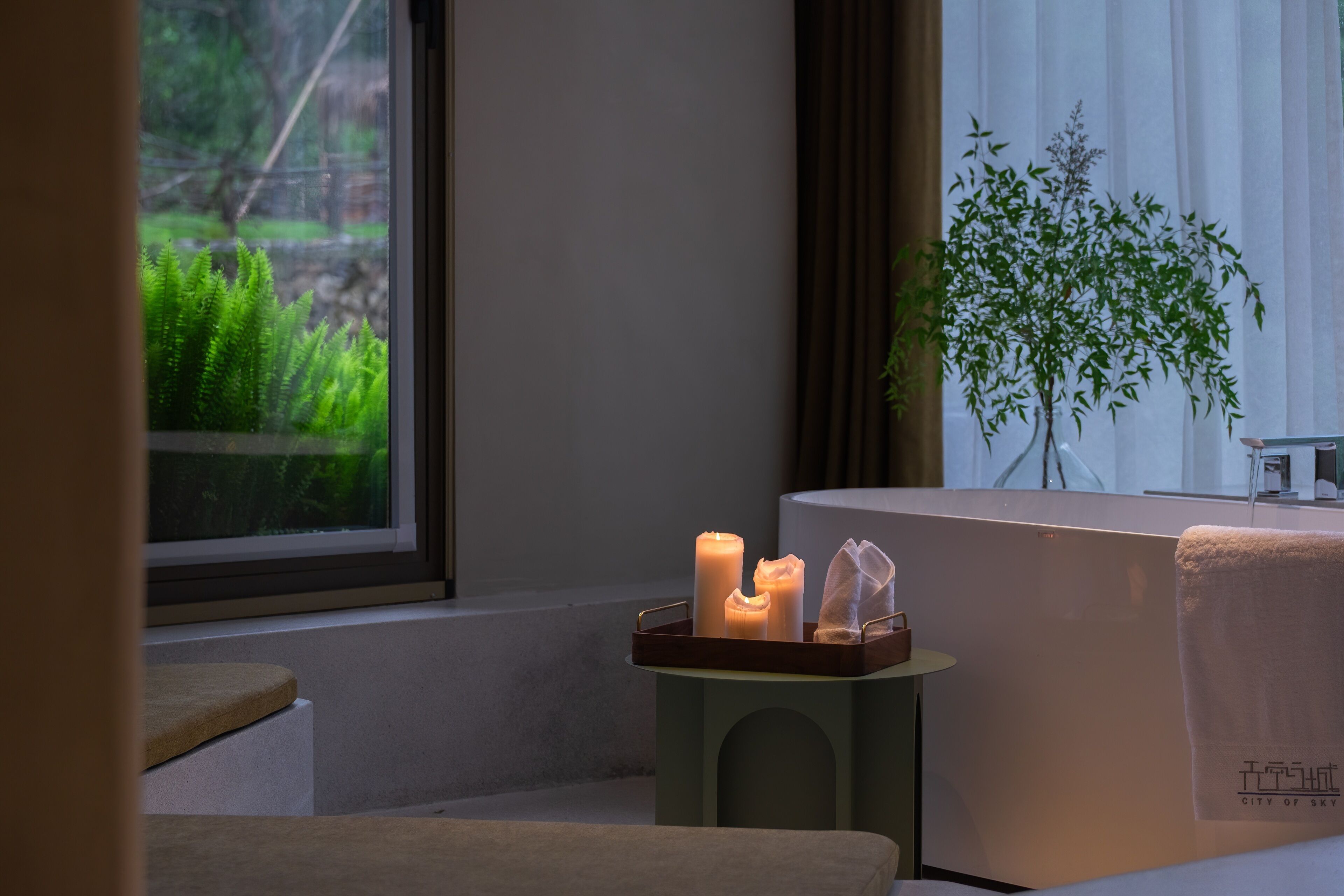 A room with a window, a small table with candles, a plant, and a towel on a chair.