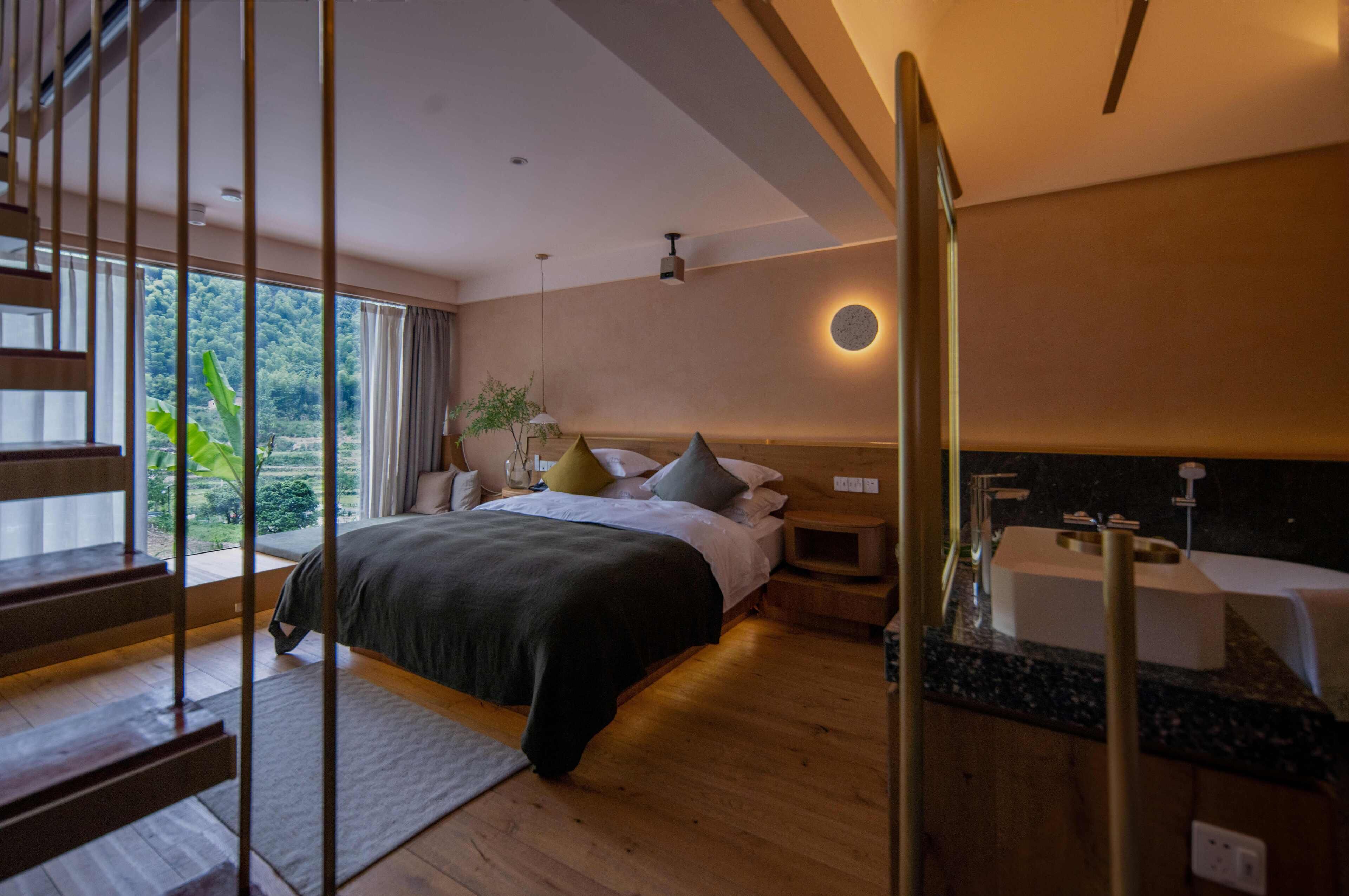 A modern bedroom with a large bed, a bedside table, a nightstand, a built-in sink, and a view of greenery outside.