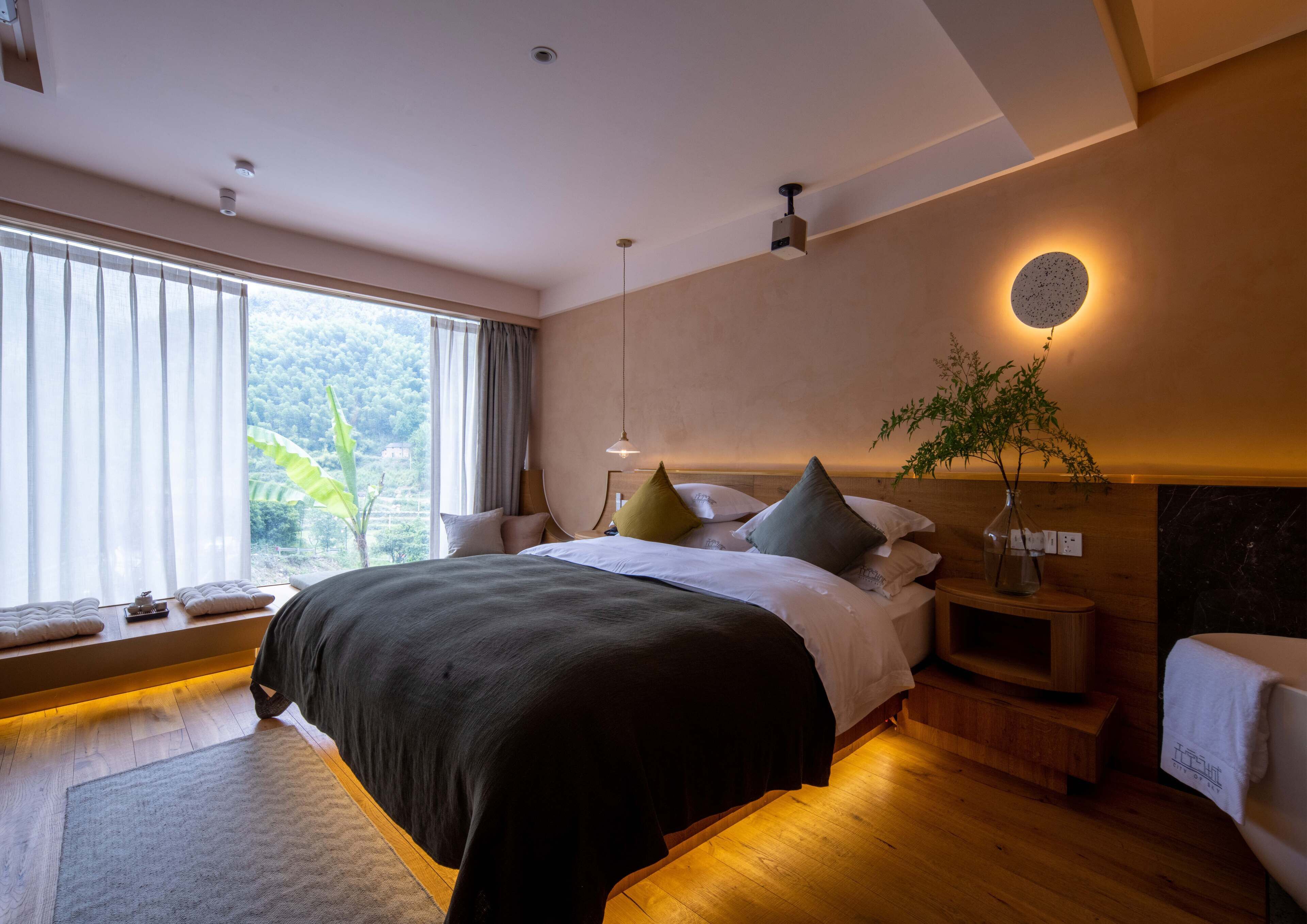 Ein modernes Schlafzimmer mit einem großen Bett, Nachttischen, einer Wandlampe und Blick auf Grünfläche vor dem Fenster.