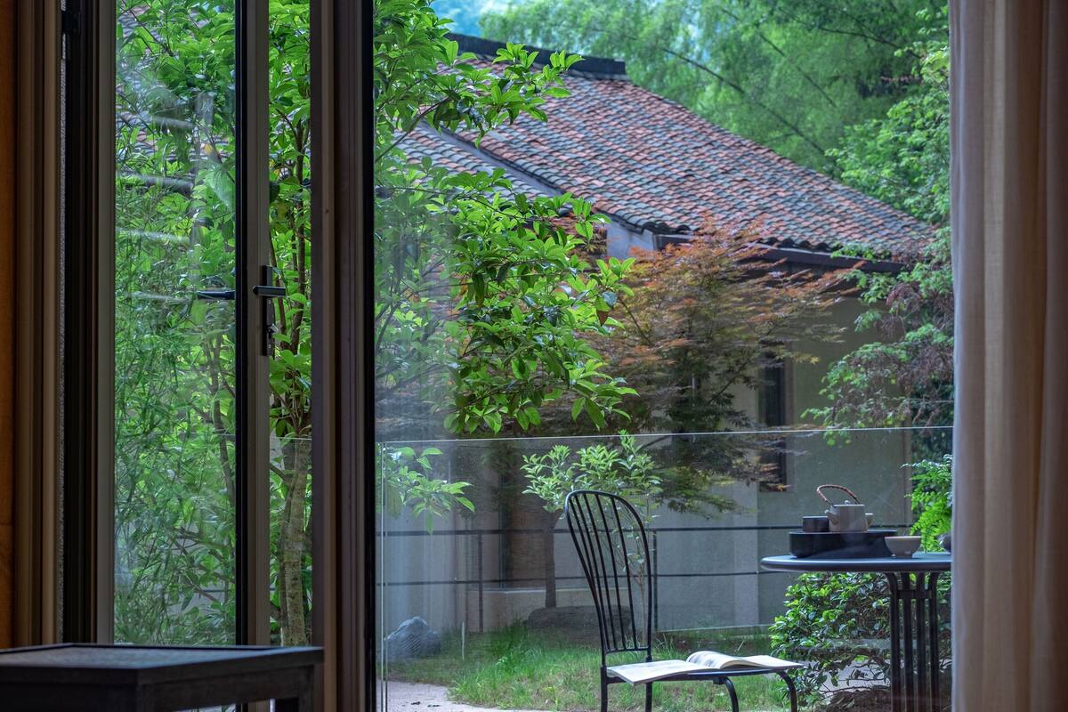 Vista a través de una ventana que muestra una habitación con una silla, una mesa, un tejado de baldosas y follaje en el exterior.
