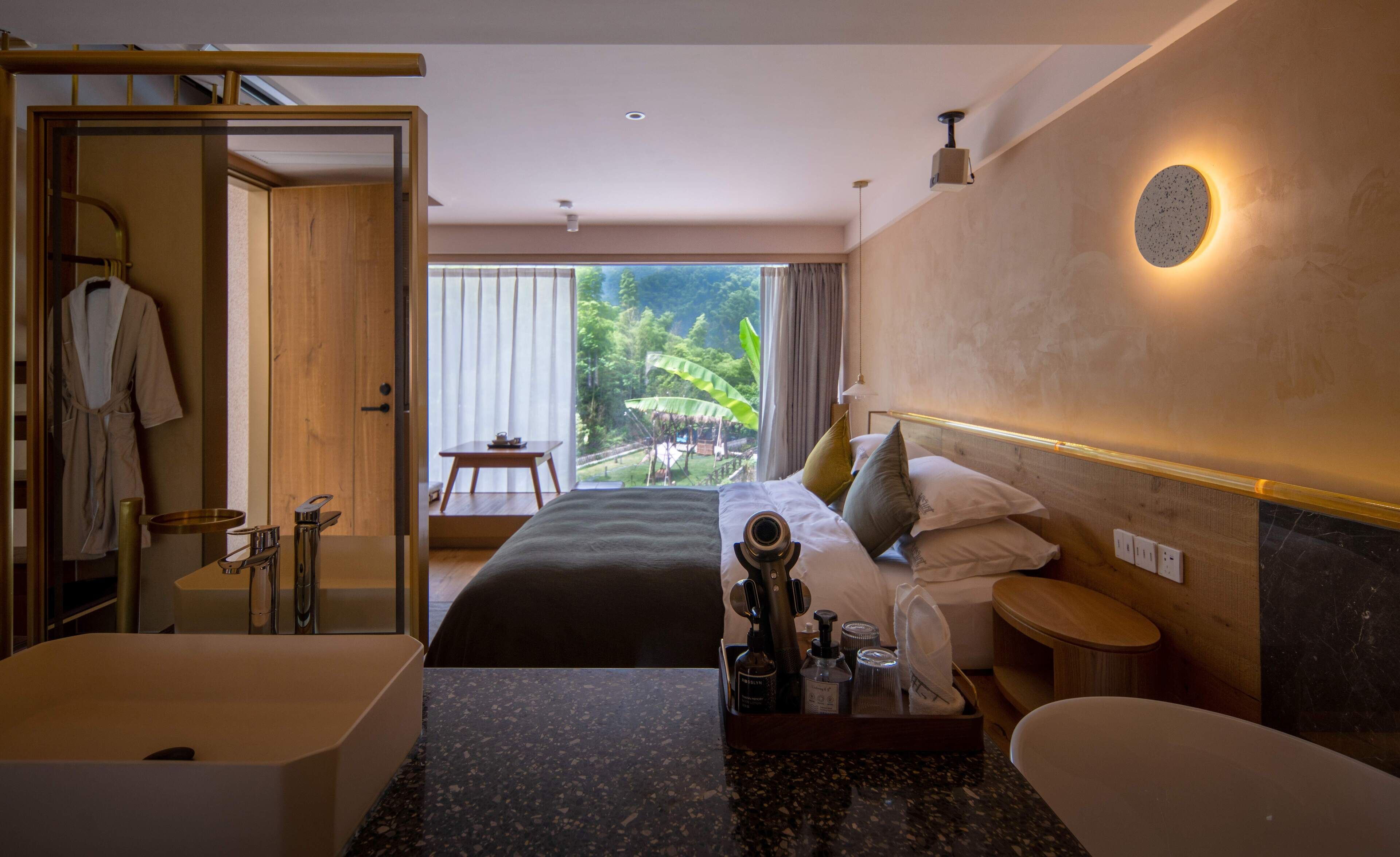 A modern hotel room with a large bed, a separate bathroom with a bathtub, a vanity with a mirror, and a view of a lush garden through the window.