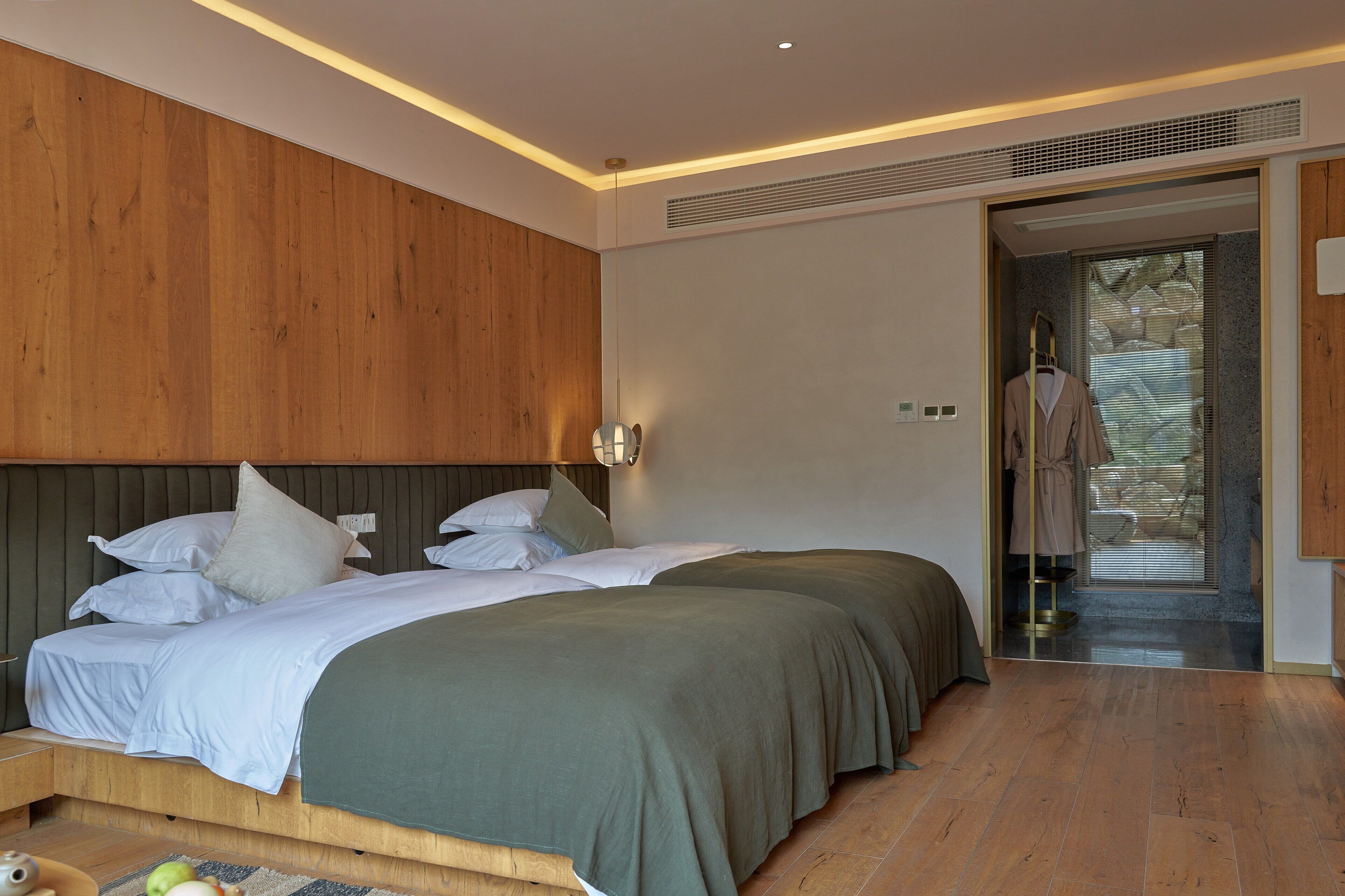 A modern hotel room with two beds, wooden paneling on the walls, a large window with blinds, and a visible bathroom door.