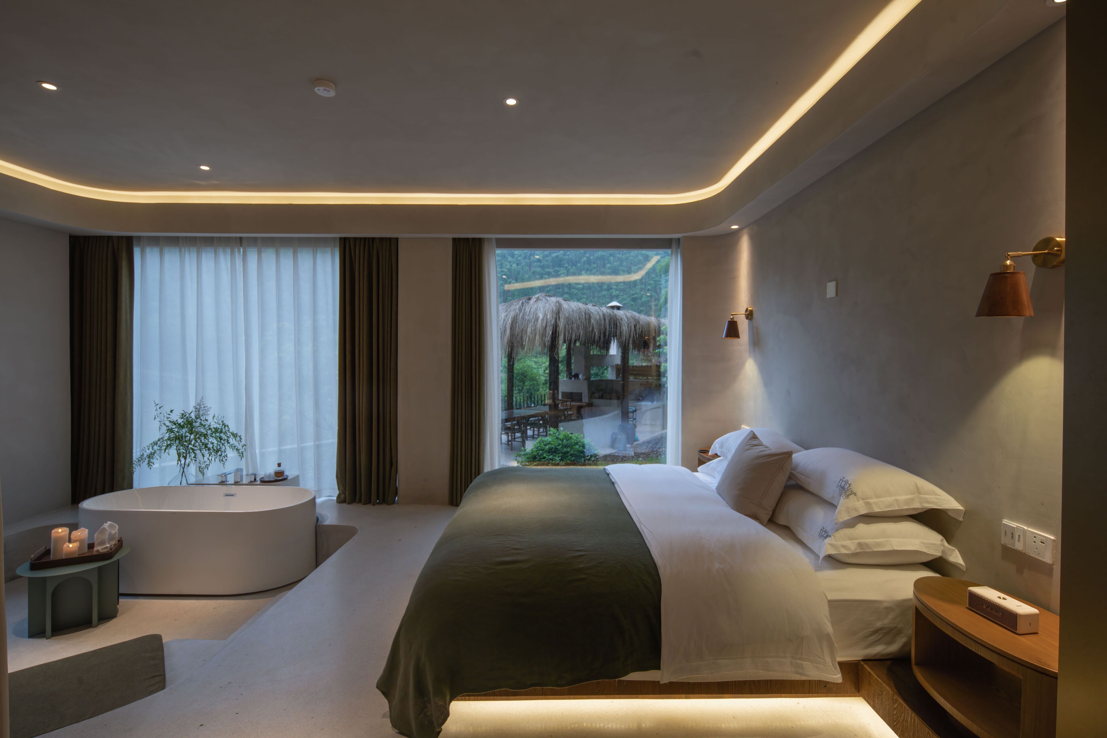 A modern bedroom with a large bed, a bathtub, and a view of a thatched-roof structure outside.