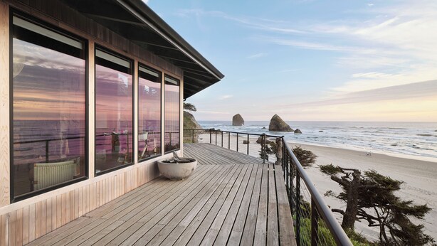 A wooden deck with large windows overlooking a beach with rock formations. - Luxury, Oceanfront, Fully Renovated, Signature MCM House in Cannon Beach (Cannon Beach)
