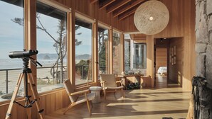A wooden interior with large windows overlooking a beach, a telescope, and a rustic stone wall. - Luxury, Oceanfront, Fully Renovated, Signature MCM House in Cannon Beach (Cannon Beach)