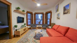 Ein gemütliches Wohnzimmer mit einem roten Sofa, einem Holztisch und zwei Sesseln. Zwei Fenster mit Fensterläden, ein Deckenventilator und ein Flachbildfernseher an der Wand sind ebenfalls vorhanden.