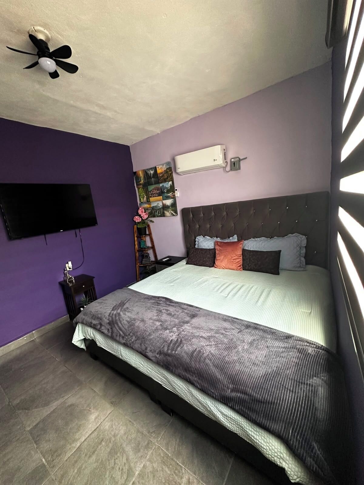 A bedroom with a bed, a ceiling fan, a mounted air conditioner, and a wall-mounted TV.