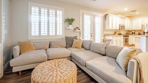 Ein Wohnzimmer mit Ecksofa, einem runden Gewebepuff und einer Küche mit weißen Schränken und einer Mikrowelle.