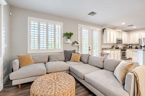 A modern living room with a sectional sofa, a round woven ottoman, and a kitchen in the background.