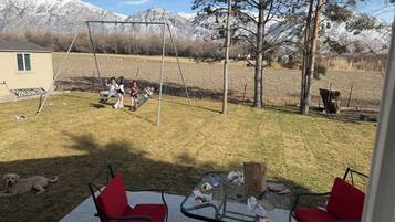 A backyard with a swing set, a glass table with red chairs, and a dog.