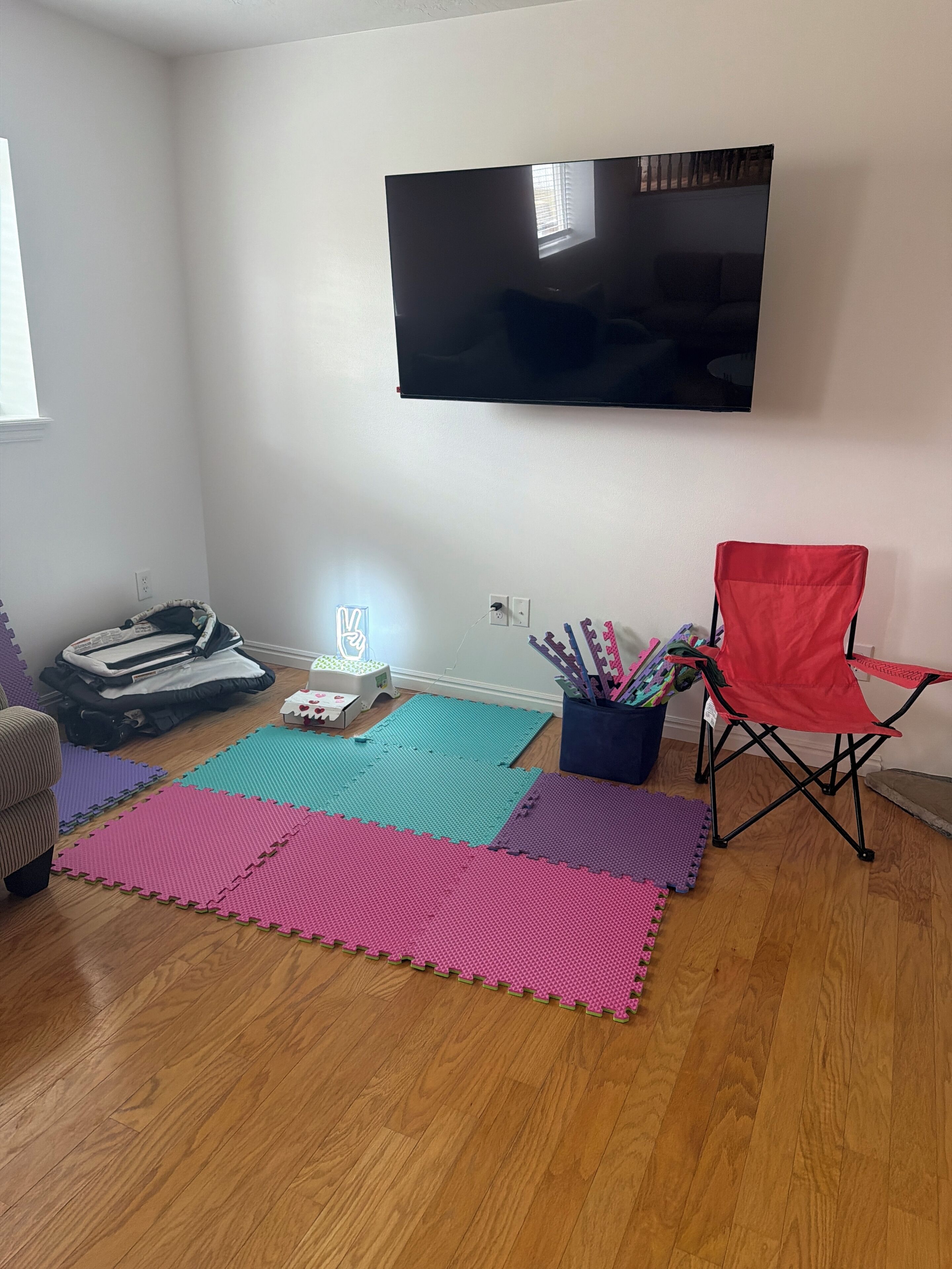 Un salón con un televisor de pantalla plana montado en la pared, una silla plegable roja y coloridos paneles de espuma entrelazados en el suelo.