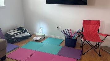 Un salón con un televisor de pantalla plana montado en la pared, una silla plegable roja y coloridos paneles de espuma entrelazados en el suelo.