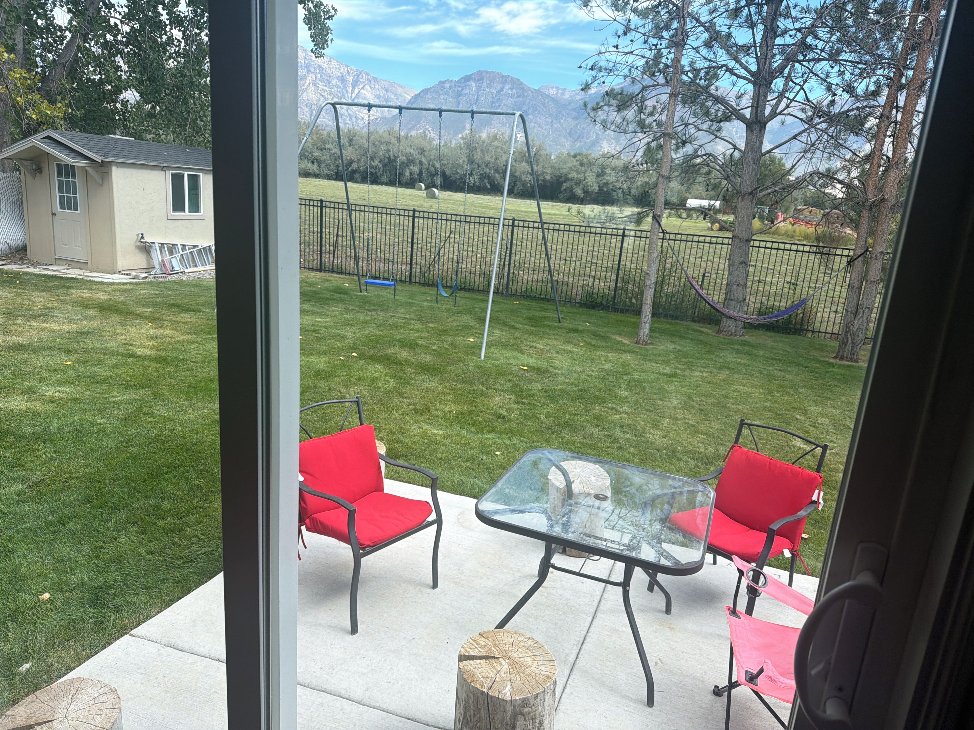 A patio with a glass table and two red chairs, a swing set, and a view of mountains.