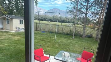 A patio with a glass table and two red chairs, a swing set, and a view of mountains.