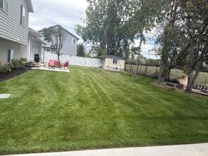 A residential backyard with a lawn, a patio with red chairs, and a swing set.