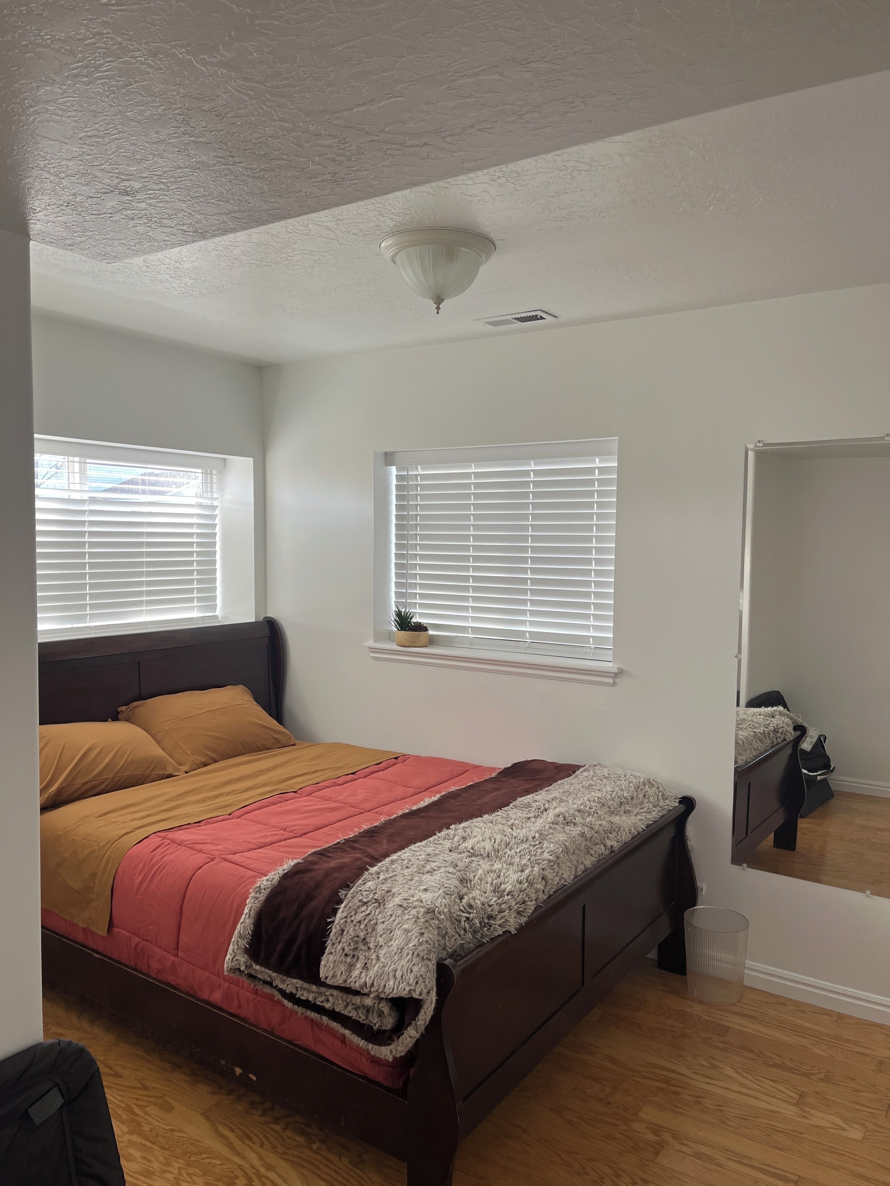 Quarto com cama, janela com persianas, luminária no teto e uma pequena planta em vaso no peitoril.