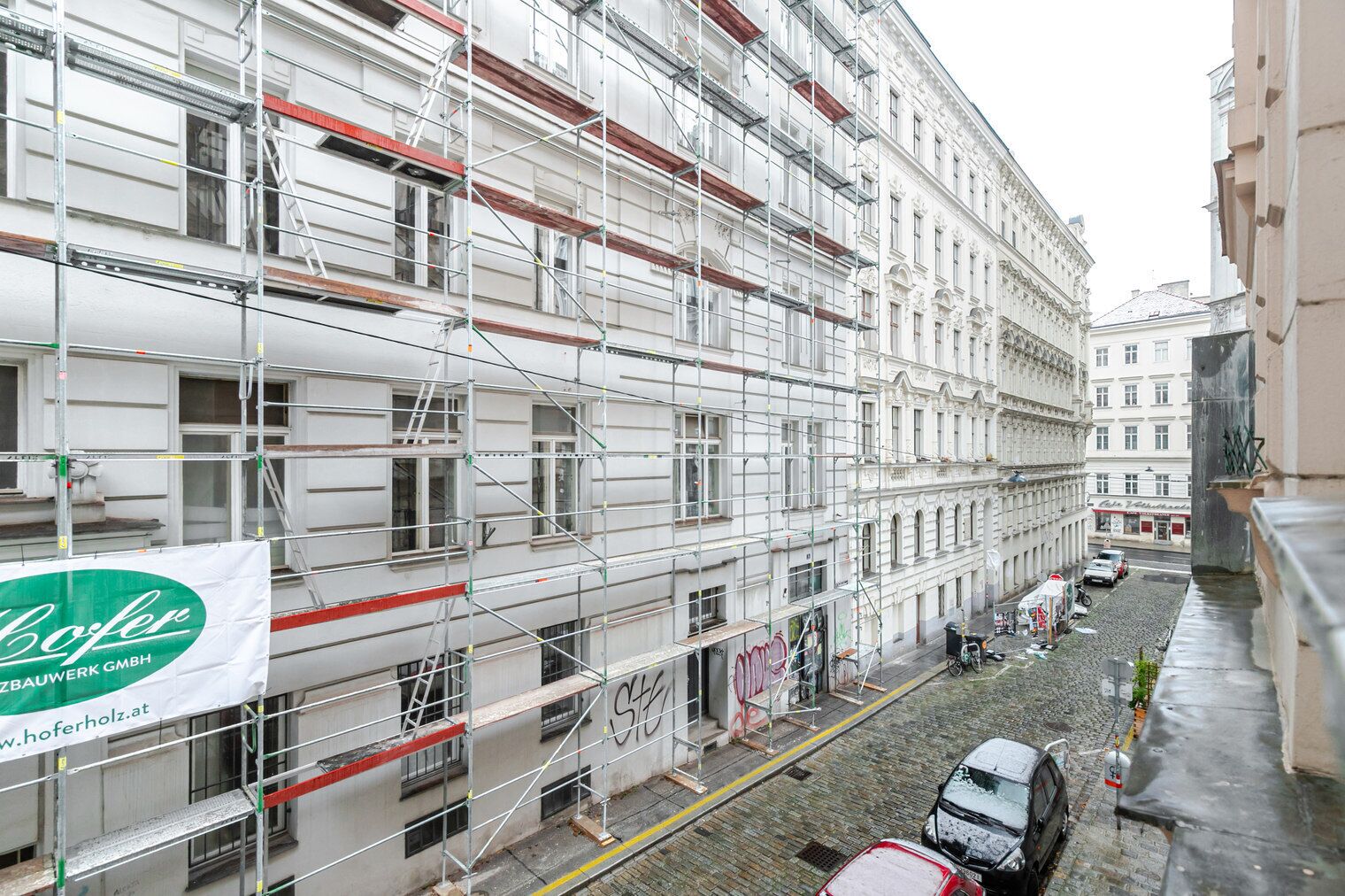 Un bâtiment en construction, avec des échafaudages, une enseigne indiquant « Hoferholz GmbH » et une rue pavée o ù sont garées des voitures.